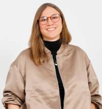 Frau mit langen braunen Haaren und runder Brille, trägt beigen Blouson über schwarzem Rollkragenpullover, lächelt.