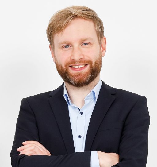 Mann mit blondem Haar und rötlichem Bart, schwarzem Blazer, hellblauem Hemd, Arme verschränkt, lächelt, weißer Hintergrund.