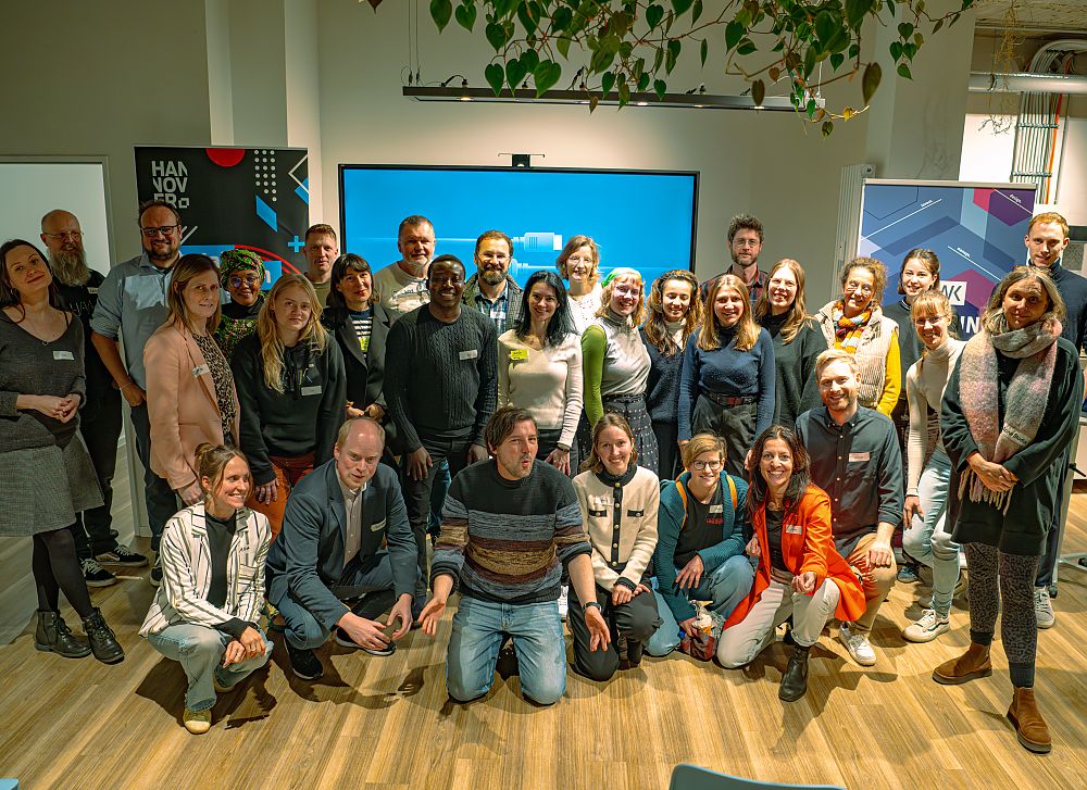 Gruppe von ca. 25 Personen posiert für ein Gruppenfoto in einem modernen Büroraum mit großem Bildschirm.