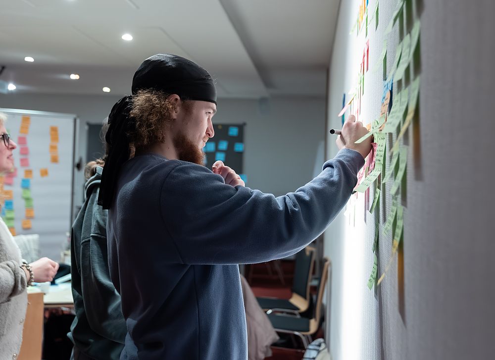 Mann mit Bart und Mütze schreibt mit Marker auf Post-its an einer Wand. Zwei Personen stehen daneben.