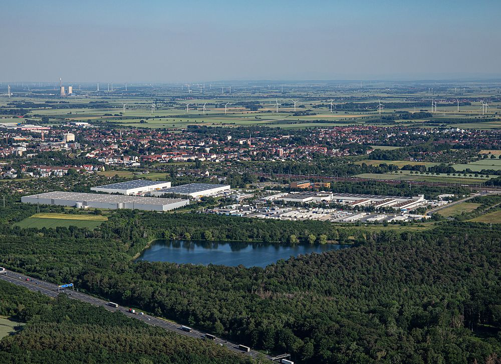 Luftaufnahme einer Stadt mit Industriegebäuden, einem See, Wäldern, Feldern und Windrädern am Horizont.