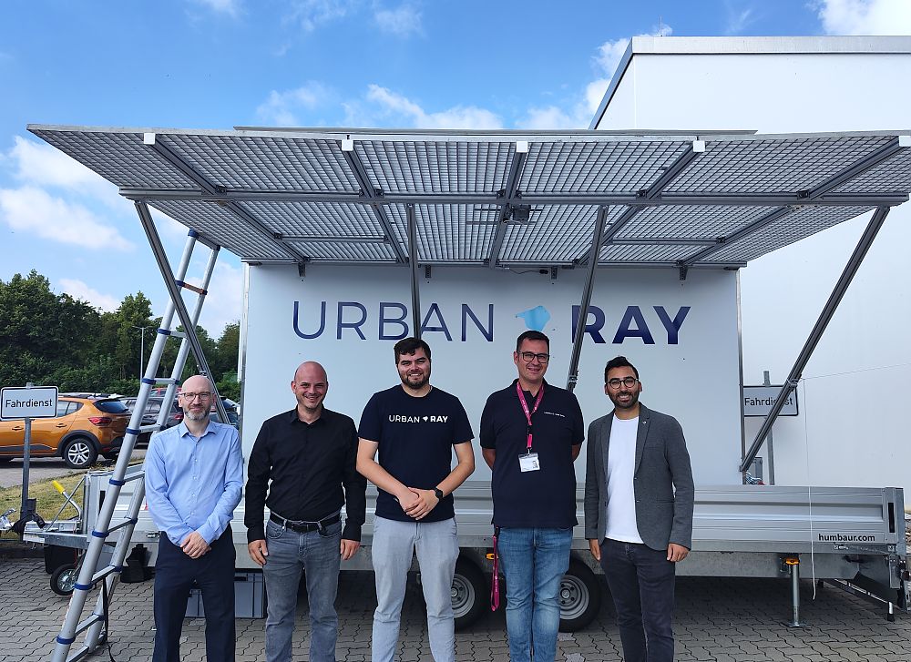 Fünf Männer stehen vor einem weißen Anhänger mit Solarpanelen und dem Logo „Urban Ray" bei bewölktem Himmel.