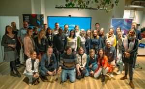 Gruppe von ca. 25 Personen posiert für ein Gruppenfoto in einem modernen Büroraum mit großem Bildschirm.
