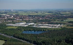 Luftaufnahme einer Stadt mit Industriegebäuden, einem See, Wäldern, Feldern und Windrädern am Horizont.