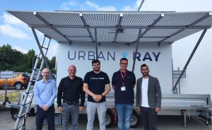 Fünf Männer stehen vor einem weißen Anhänger mit Solarpanelen und dem Logo „Urban Ray" bei bewölktem Himmel.