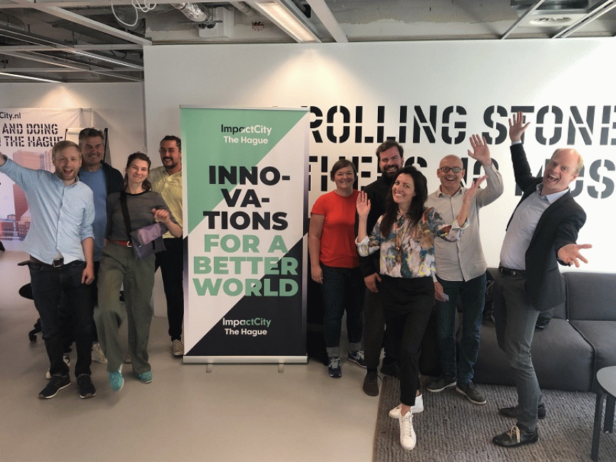 Gruppe von 9 Personen posiert vor einem ImpactCity-Banner mit Aufschrift „Innovations for a Better World" in The Hague.